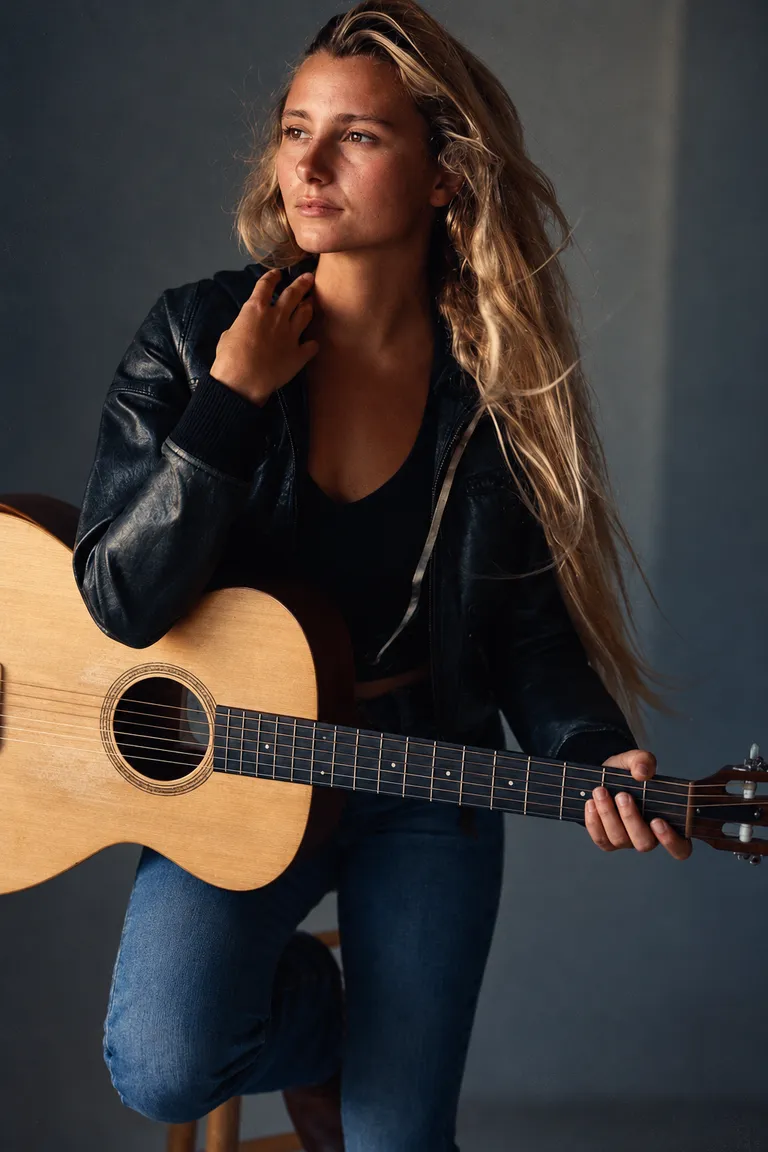 Lucy Spotts seated in a studio, wearing a leather jacket and holding an acoustic guitar.