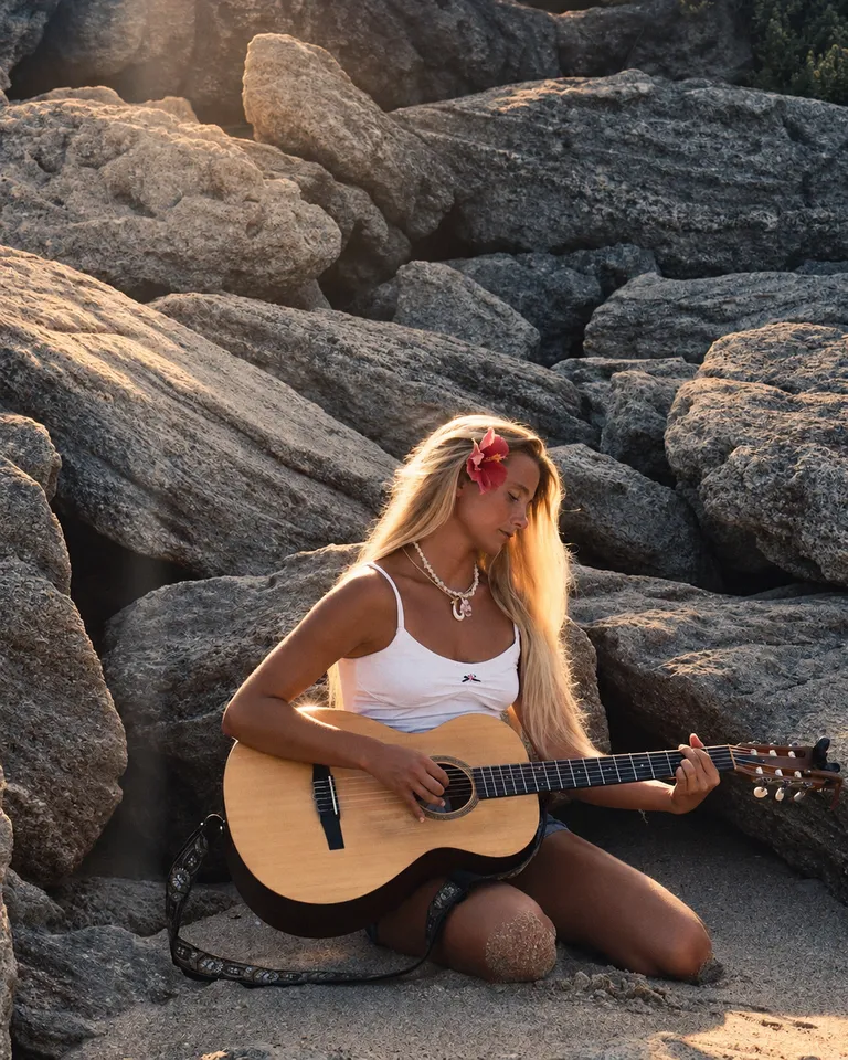 Lucy Spotts with an acoustic guitar among beach rocks.