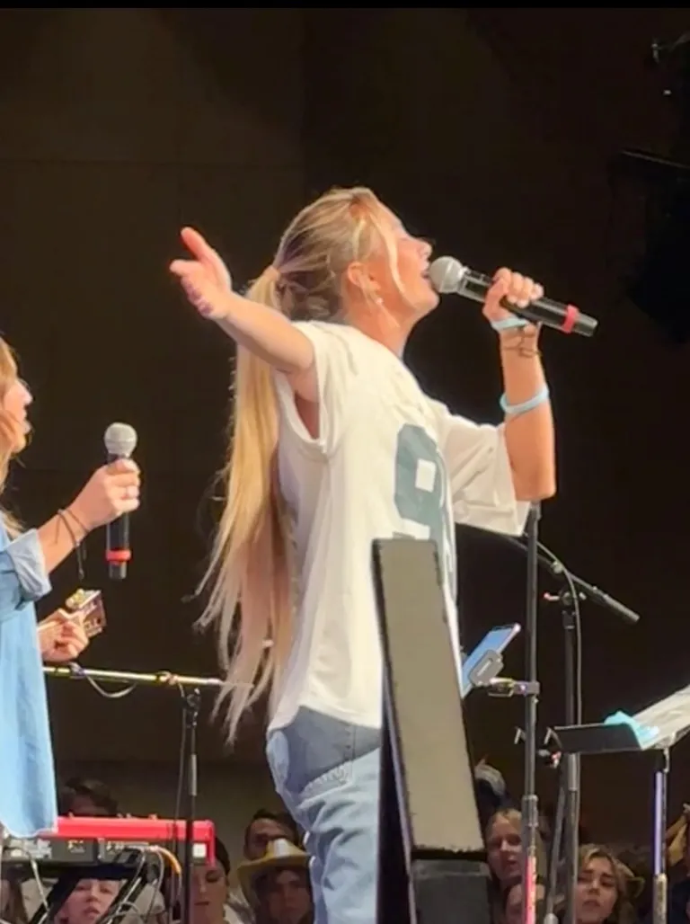 Lucy Spotts leading worship on stage, hair in a ponytail, wearing a white t-shirt, one hand raised and a microphone in the other.