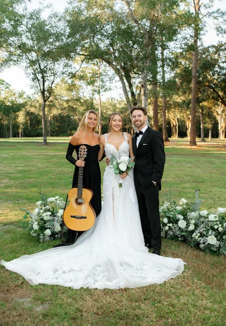 Lucy Spotts in a green dress with a guitar beside the bride and groom on a lawn.