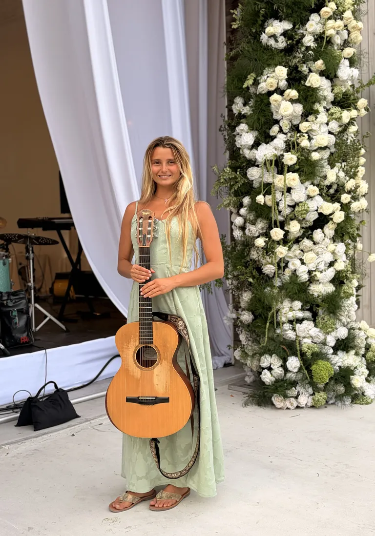 Lucy Spotts in a green dress with a guitar indoors, in front of a white flower wall.