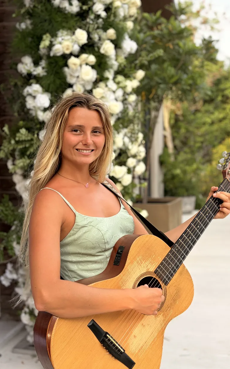 Lucy Spotts close up in a green dress with a guitar, flowers behind her.