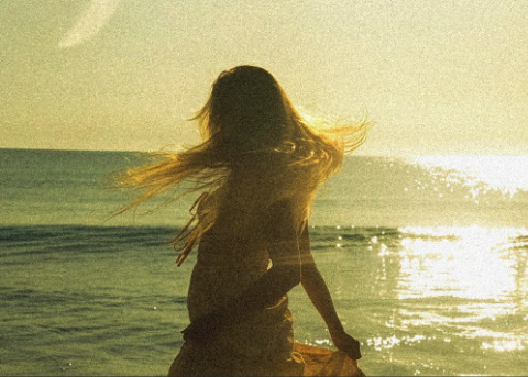 Lucy Spotts in a floral dress playing acoustic guitar on the beach.